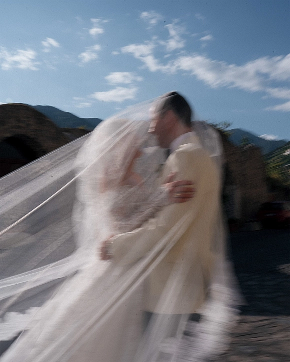 Wedding kiss portrait under a windblown veil as the bride and groom embrace, her dress flowing in a rocky mountain landscape beneath blue sky clouds
