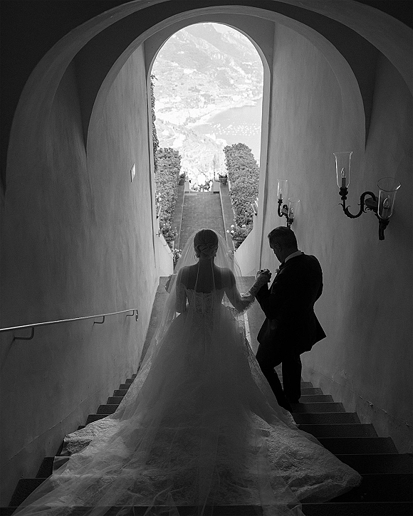 Bridal entrance as bride walking down stairs with her father, long train and veil flowing by an arched stone doorway with sconces