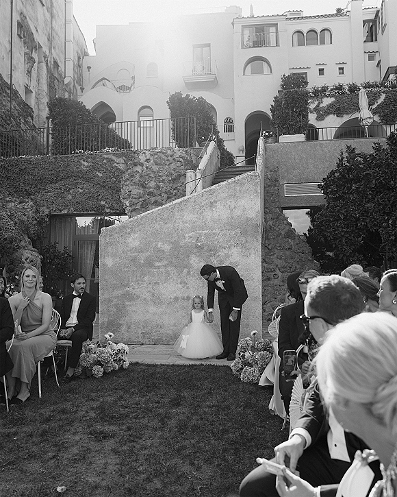 Ceremony moment as a tuxedoed groom stands beside a flower girl walking down aisle, with guests in an ivy-lined courtyard venue