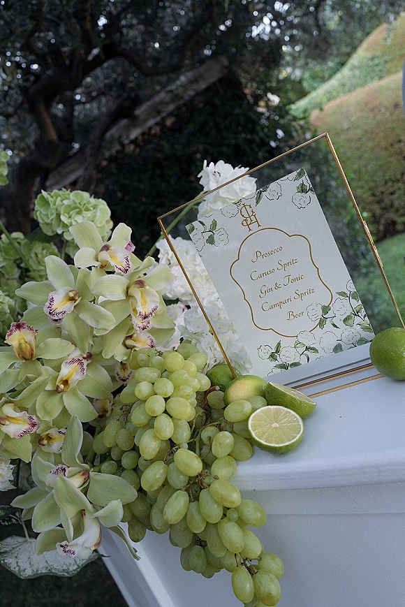 Wedding bar menu in a gold frame with orchids, hydrangea, green grapes and limes on display at an outdoor garden bar setup