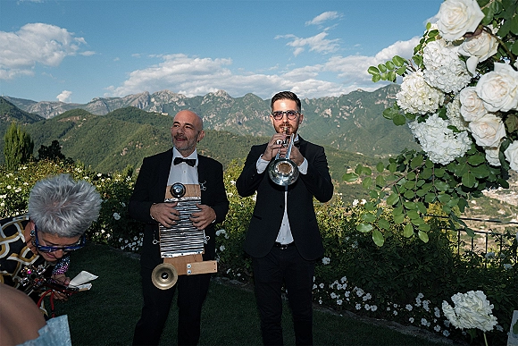 Wedding musicians in black tuxedos play trumpet and washboard under a white floral arch on a garden terrace with mountain views