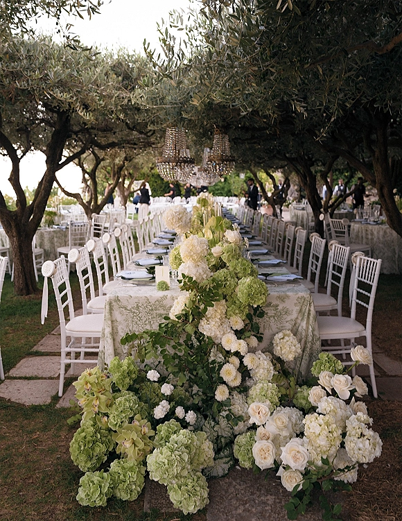 Reception tablescape at an outdoor wedding reception with a long banquet table, green-and-white floral runner, and chandeliers under olive trees