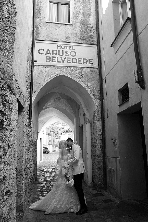 Wedding kiss portrait of bride and groom kissing beneath a stone archway, bridal veil draped over faces in a cobblestone alley
