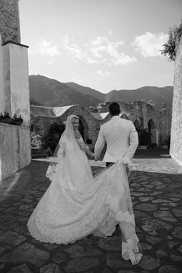 Couple portrait of newlyweds holding hands walking away, bride looking back as her long veil flows through a stone courtyard ruins backdrop