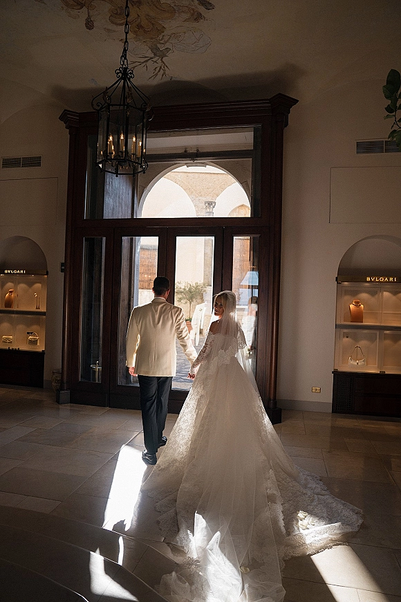 Wedding couple portrait of bride and groom holding hands, walking through arched glass doors as she looks back, veil trailing in sunlit lobby