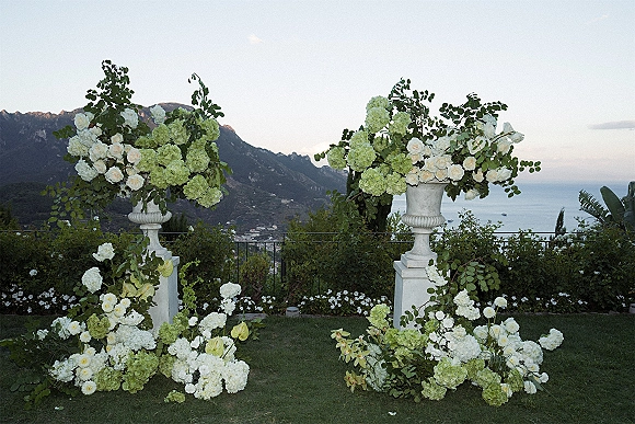 Wedding altar flowers in twin urn vases with white roses and hydrangeas on pedestal stands, set on a terrace with ocean and mountain views