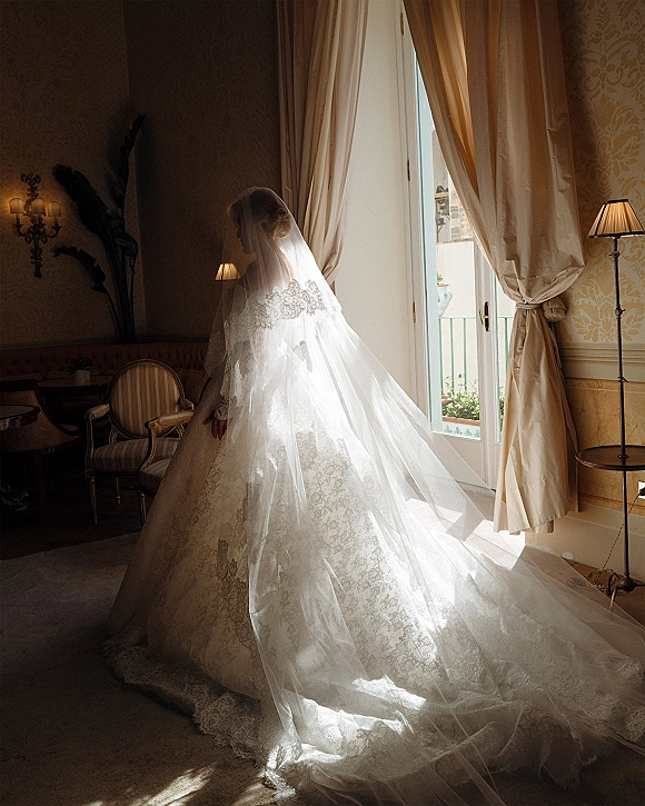 Bridal portrait of a bride by window in a hotel suite, wearing a cathedral veil and lace wedding dress with a long train in soft light