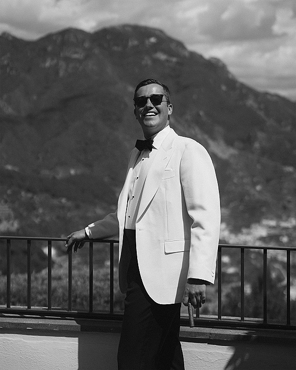 Groom portrait in a white tuxedo jacket with black bow tie, sunglasses and cigar, leaning on balcony railing with mountains behind