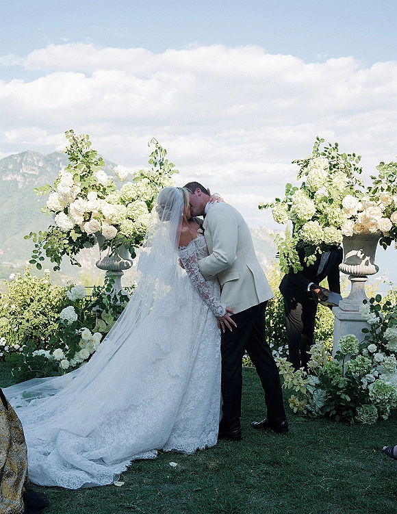Wedding kiss portrait of the couple sharing a ceremony kiss moment, bride in lace dress and cathedral veil with mountain sky behind