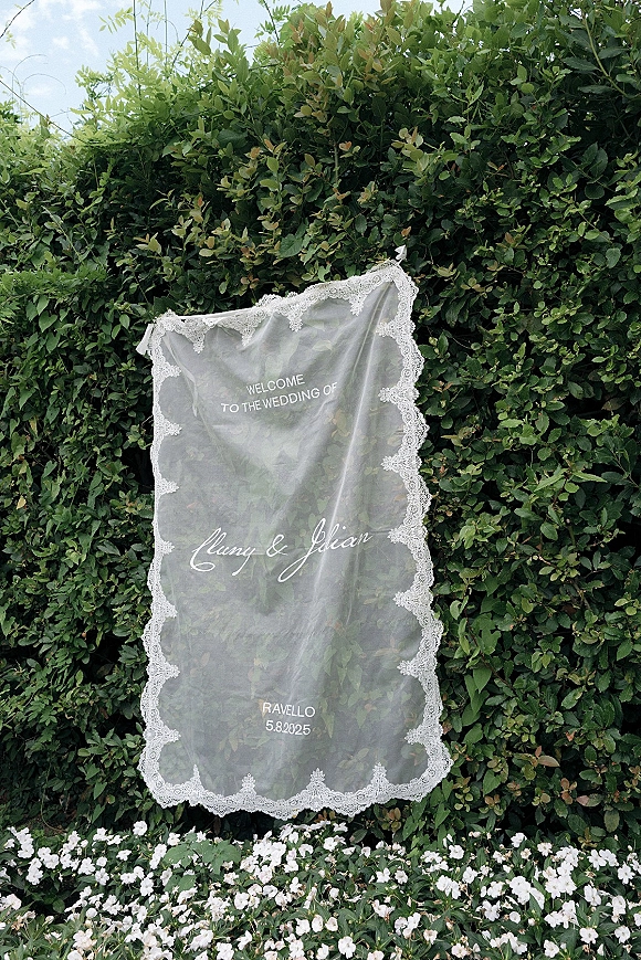 Wedding welcome sign, acrylic wedding welcome sign with lace trim and white lettering set against a green hedge with white flowers outdoors