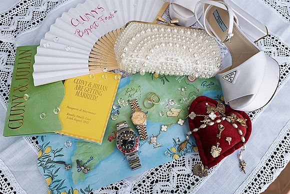 Invitation suite wedding invitation flat lay with envelope, rings, pearl clutch, white bridal heels, and watches on lace tablecloth and map print