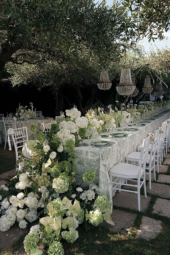 Reception tablescape with an outdoor banquet table, floral garland, taper candles, and crystal chandeliers hanging in olive trees overhead