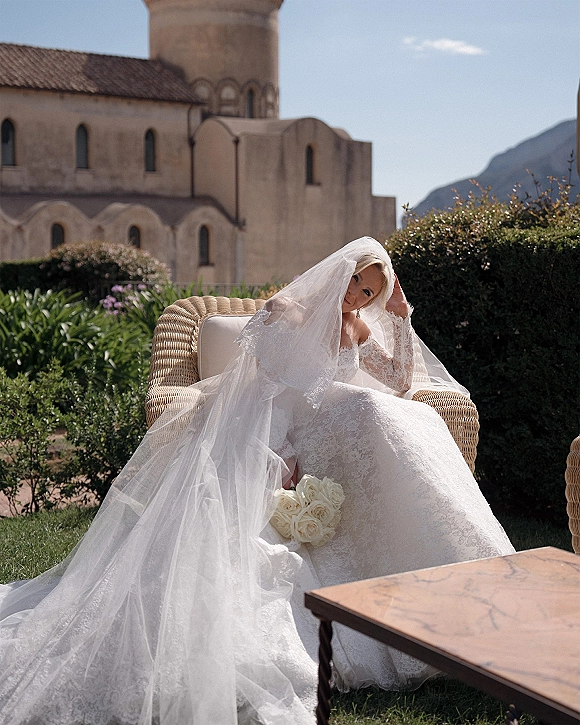 Bridal portrait of a bride seated on a wicker chair in a lace-sleeve gown, holding a white rose bouquet with mountains behind her