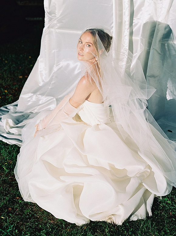 Bridal portrait of a bride in a strapless wedding dress with veil over her face, pearl jewelry and gloves, seated on grass by a draped white backdrop