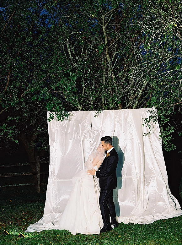 Wedding couple portrait of bride and groom embrace under flash, her veil and gown against a white fabric backdrop on a lawn at night