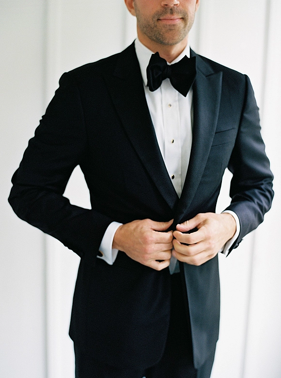 Groom portrait in a tuxedo with a black bow tie and crisp white dress shirt, standing against a clean white wall indoors