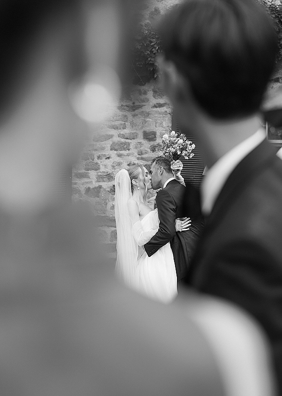 Wedding kiss as the bride and groom kissing, her veil and bouquet raised, framed by wedding guests with a rustic stone wall behind