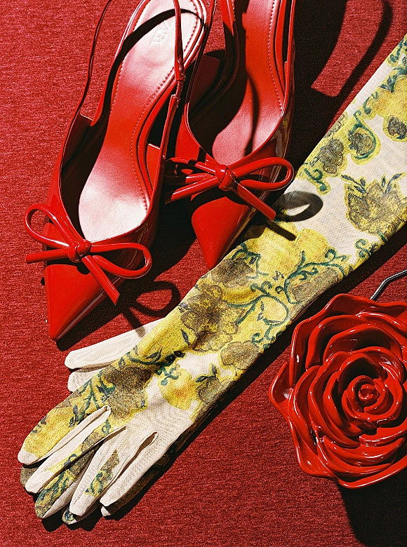Bridal shoes flatlay featuring red wedding heels with bow detail, floral gloves and a red rose accessory on red fabric backdrop