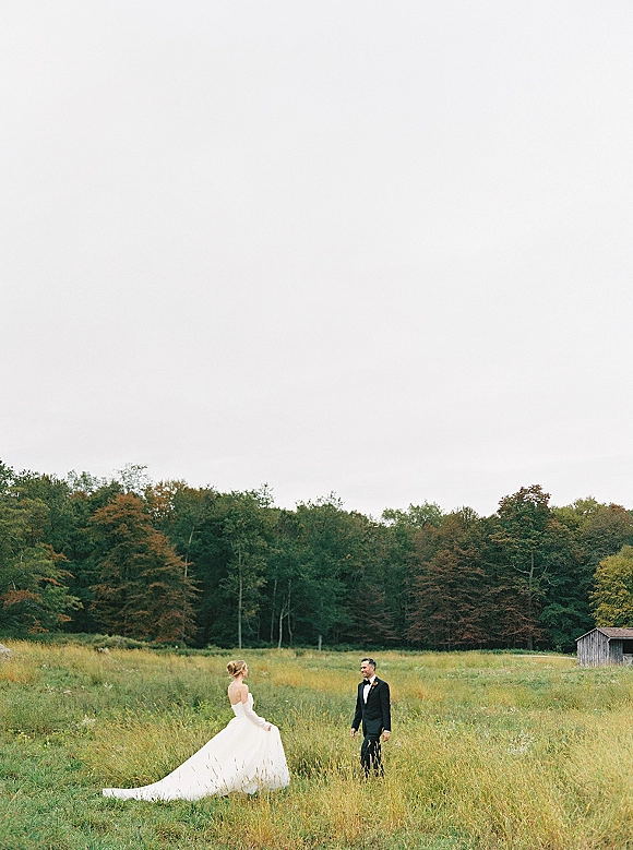 Couple portrait of bride and groom in field, facing each other as she lifts her long train in tall grass near a rustic shed.