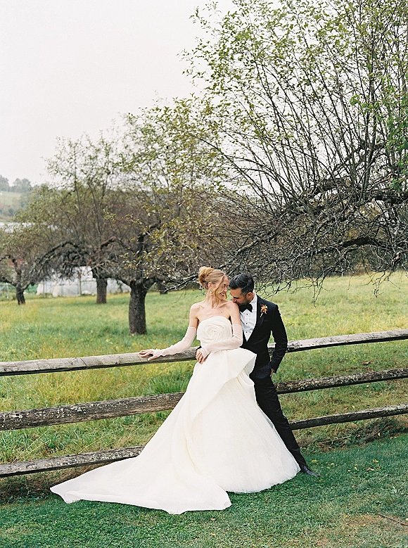 Couple portrait of bride in strapless gown with long train and opera gloves as groom embraces her by a wood fence in an orchard field