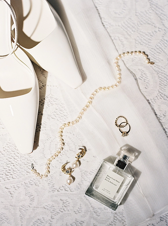 Bridal details flatlay with white wedding heels, pearl necklace and earrings, rings, perfume bottle, and veil on lace background