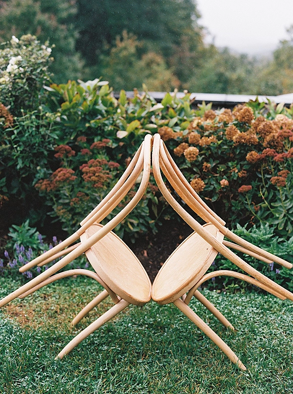 Wedding ceremony chairs in natural wood accents set on a green lawn, backed by garden greenery and flowering shrubs outdoors