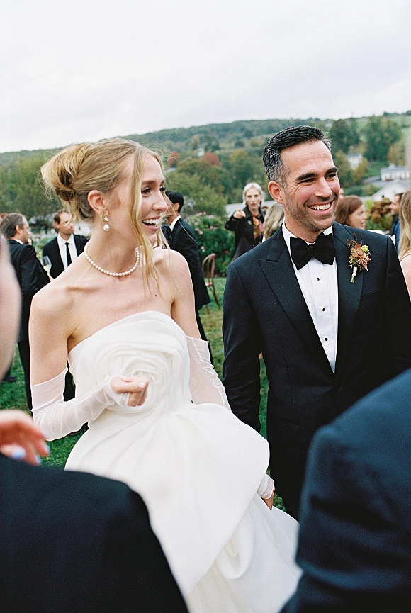 Wedding couple candid, bride and groom laughing as they walk past guests on an outdoor lawn with hills and trees under a cloudy sky
