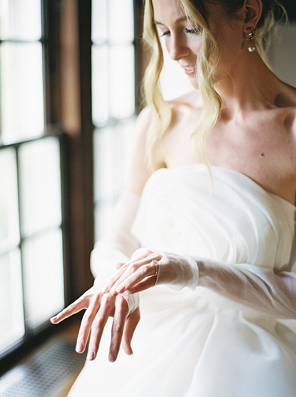 Bridal portrait of bride adjusting wedding ring in sheer gloves, wearing a strapless dress and veil, softly lit by window panes indoors