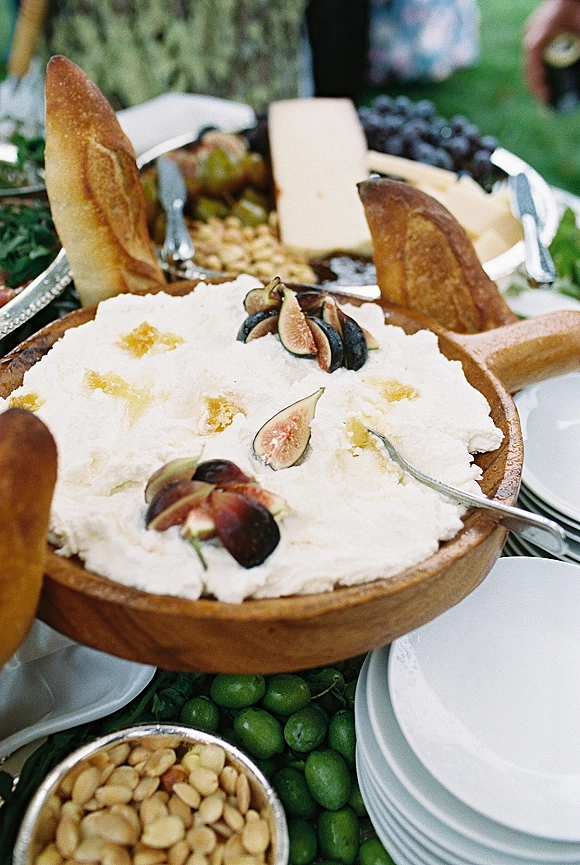 Wedding charcuterie board, wedding appetizer spread with cheese, crostini, figs, grapes and olives on a lawn with greenery and guests’ hands