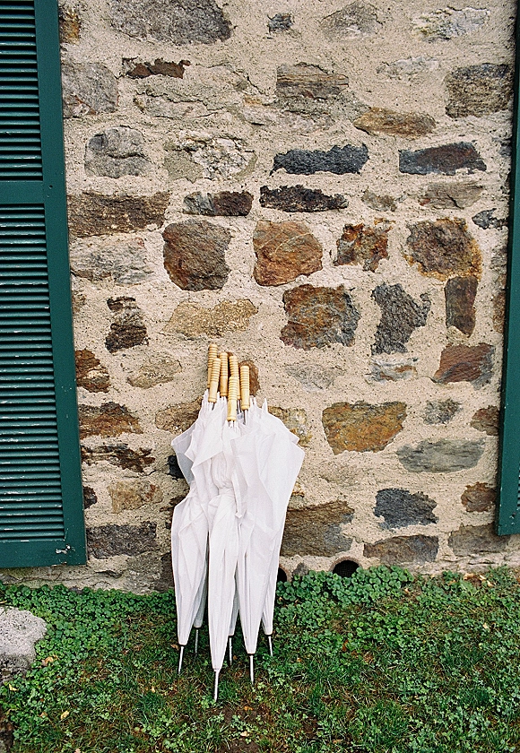 Wedding umbrellas with wooden handles lined up on grass beside a stone wall with green shutters for a rainy outdoor ceremony plan