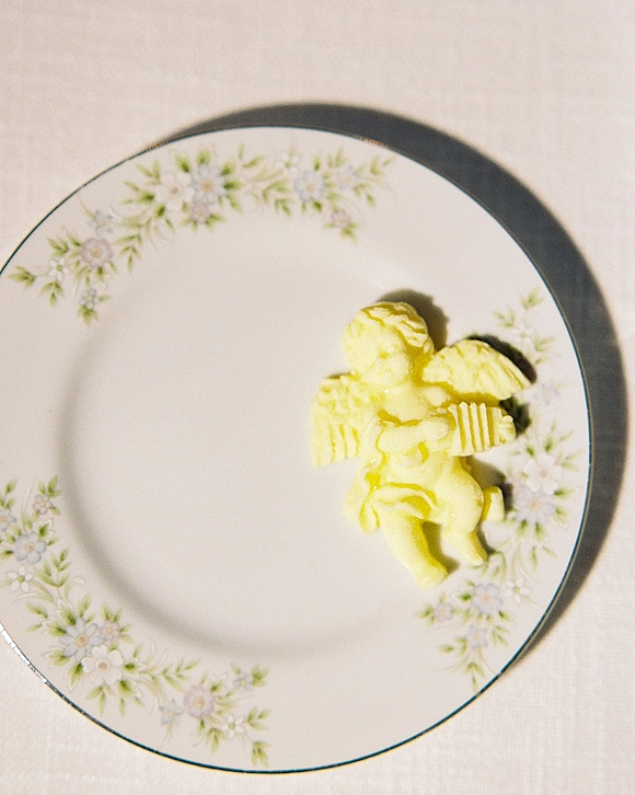 Wedding butter sculpture shaped like a butter cupid on a floral china plate, styled as a whimsical tablescape detail on a white tablecloth