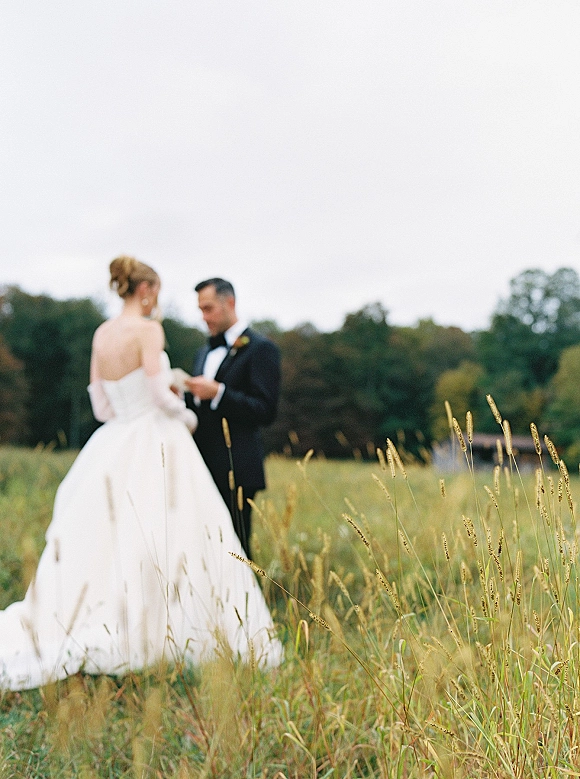 Wedding vows as groom reads from paper while bride listens in a meadow field, her strapless gown and his tuxedo under overcast sky