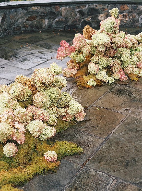 Wedding floral installation with hydrangea ceremony flowers and moss base arranged on a stone patio beside a stone wall