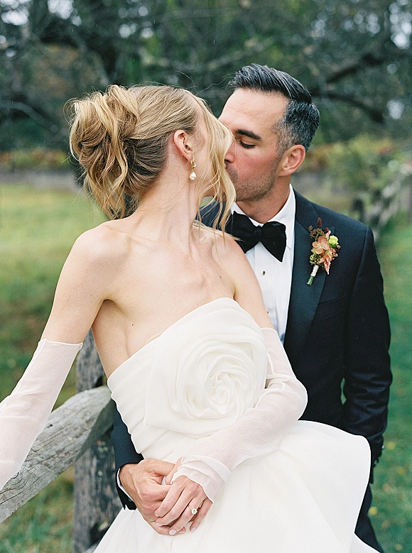 Wedding kiss portrait of bride and groom kissing as he holds her, her strapless rosette gown and pearl earrings by trees and a wooden fence