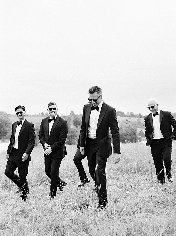 Groomsmen portrait of a group in black tuxedos and sunglasses walking together across a grassy field with rolling hills and open sky