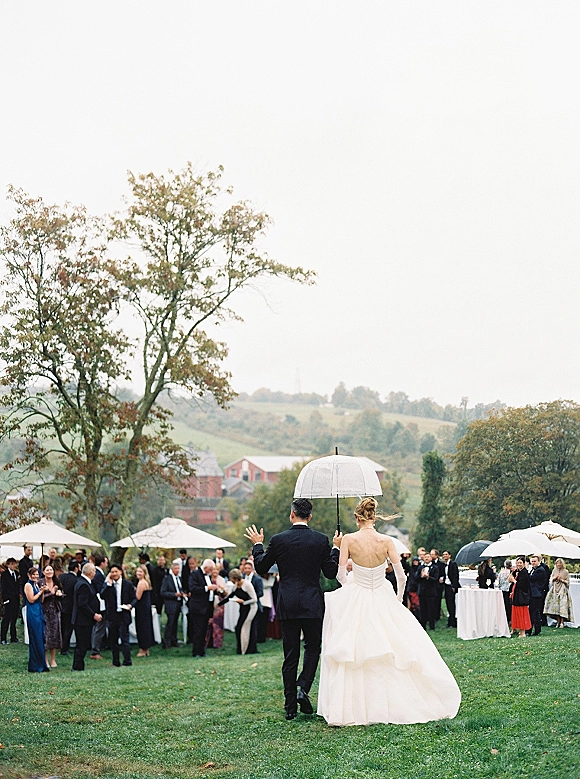 Wedding reception entrance as bride and groom walk away under a clear umbrella, guests applauding on a lawn with barn and hills behind