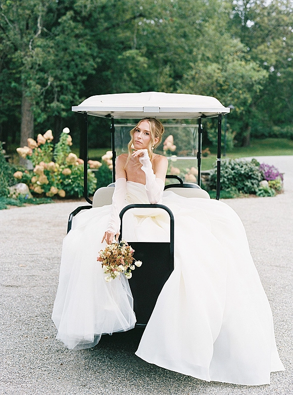 Bridal portrait of a bride on golf cart, holding a bouquet in a strapless gown and opera gloves on a gravel driveway by hydrangeas