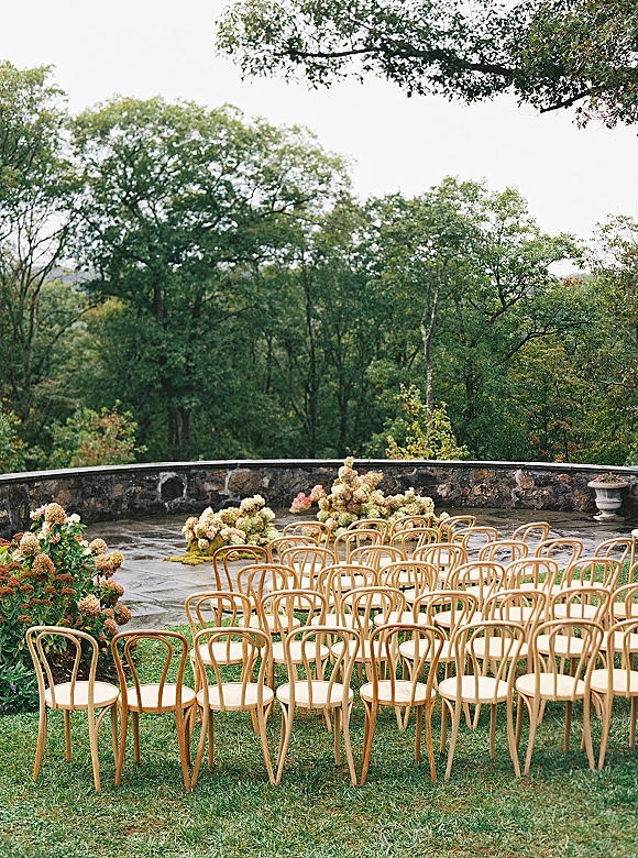 Ceremony seating with outdoor wedding ceremony seating of wood chairs arranged in a curve, lined with hydrangea florals on a stone terrace lawn