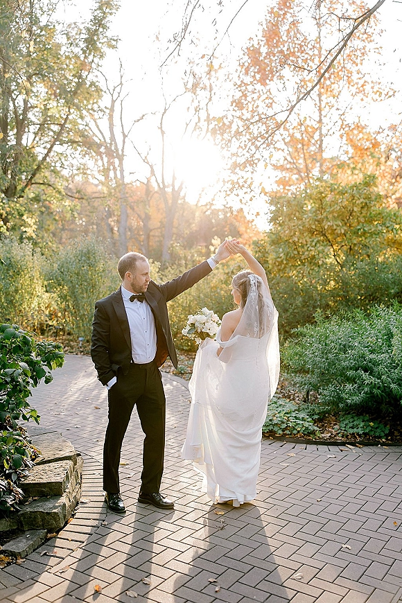 Couple portrait of bride and groom dancing as he spins her on a sunlit garden path, her veil and bouquet flowing in autumn foliage
