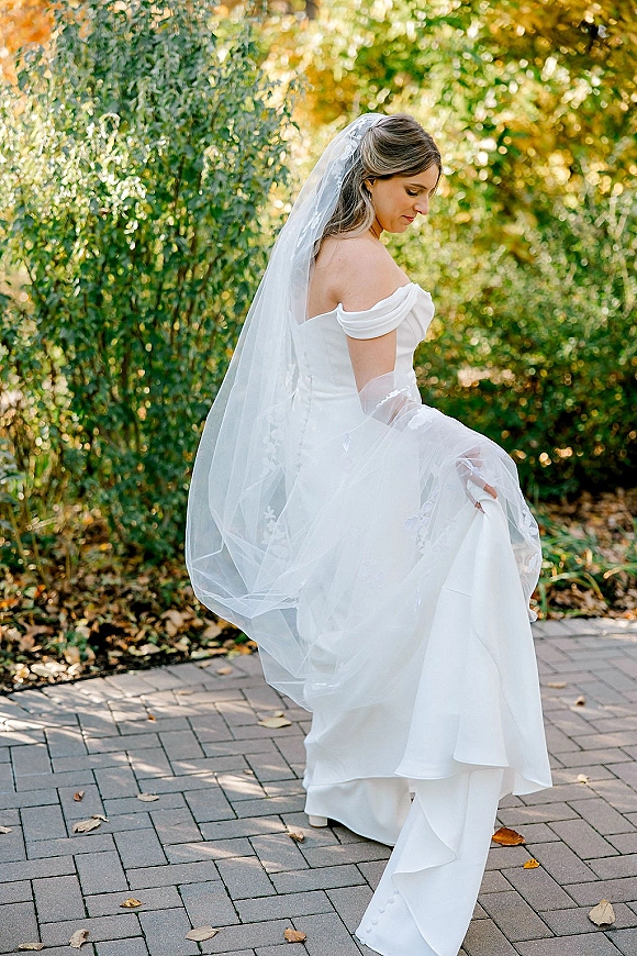 Bridal portrait of a bride looking down, holding her off-the-shoulder dress hem as her lace veil and long train trail on a brick walkway