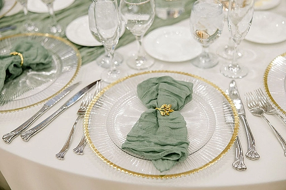 Reception tablescape wedding place setting with sage green napkins in gold rings, clear glass chargers, white plates, and wine glasses on white linens