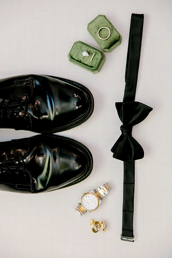 Wedding rings in a velvet ring box with black bow tie, dress shoes, watch and cufflinks arranged on white fabric flatlay
