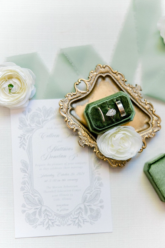 Wedding rings arranged in velvet ring box styling on an ornate gold tray with invitations and white flowers over soft white linen backdrop