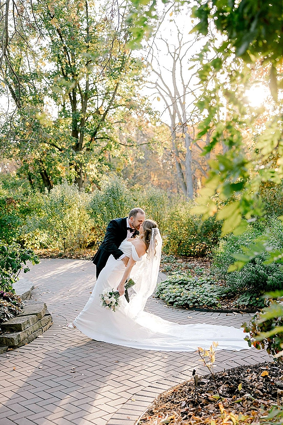 Wedding kiss portrait of groom dipping the bride, her cathedral veil and long train flowing on a sunlit brick garden path