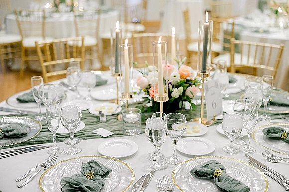 Reception tablescape with wedding table centerpieces, taper candles, sage green napkins, and floral greenery centerpiece on white linens with gold chiavari chairs