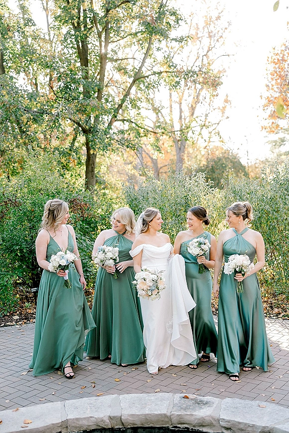 Bridesmaids photo of a bride in a veil surrounded by bridesmaids in sage green dresses holding white rose bouquets on a sunny garden path