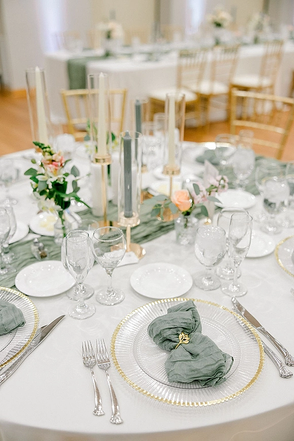 Reception tablescape with wedding table setting of sage green napkins, gold rings, taper candles in cylinder vases, and floral greenery garland under white draping