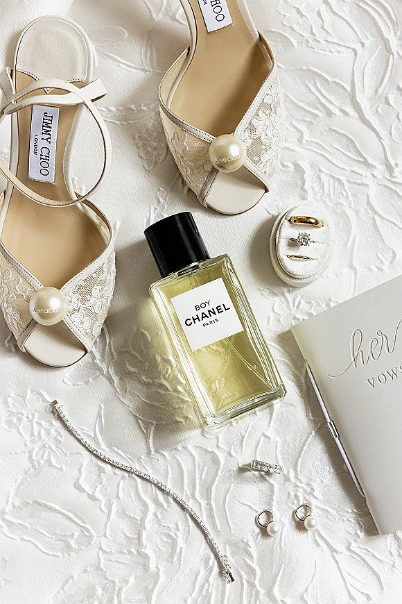 Wedding flat lay of bridal details featuring lace heels with pearl accents, perfume bottle, ring box, vow book, and bands on white fabric