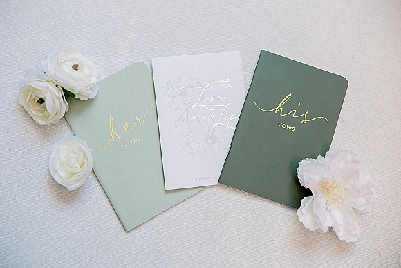 Wedding vow books with gold foil lettering in a his and her set, styled as a flat lay with white flowers on linen fabric backdrop