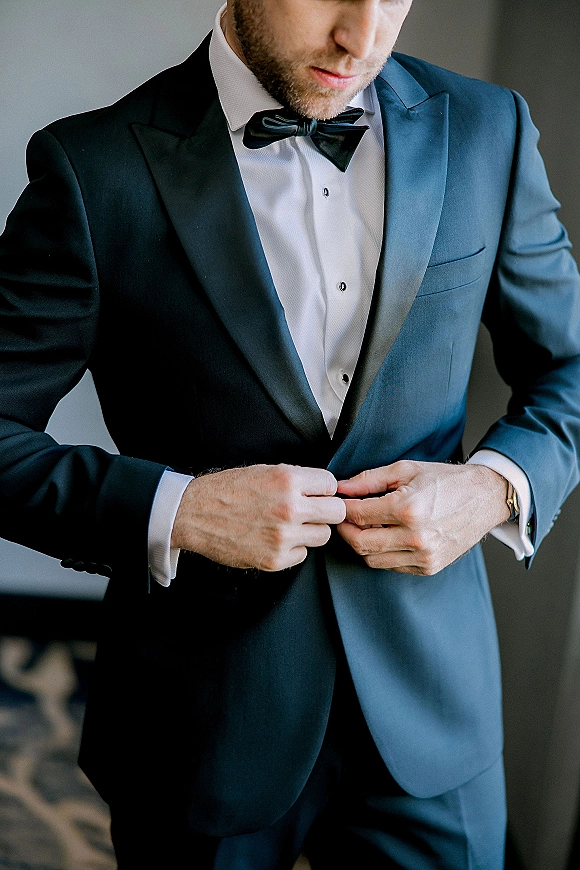 Groom getting ready, buttoning a navy tuxedo jacket with satin lapel and black bow tie in soft window light against a neutral wall